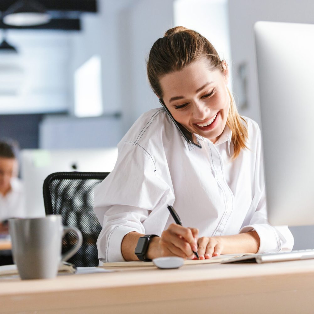 Cheerful young businesswoman sitting at her workplace at the office, talking on mobile phone, taking notes